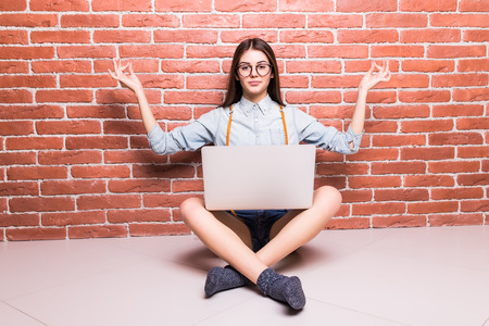 Beautiful young dark-haired girl in casual clothes posing with laptop on sholders, hands up, sitting cross-legged against brick wallの写真素材