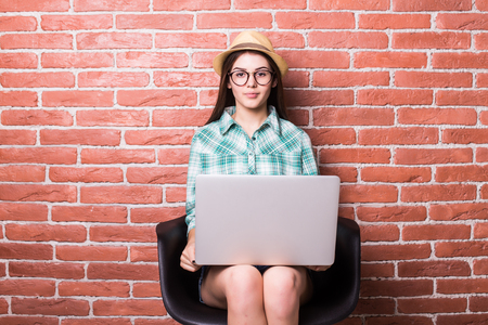 Beautiful young woman in casual wear is using a laptop and smiling, sitting against red brick wallの写真素材
