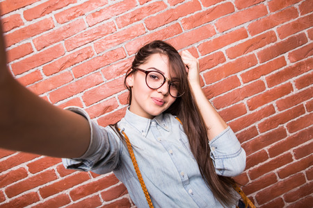 Beautiful young dark-haired girl in casual clothes and eyeglasses posing, smiling and making selfie, standing against brick wallの写真素材