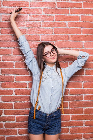 Beautiful young woman in headphones is listening to music using a smart phone, up hand and smiling, against red brick wallの写真素材