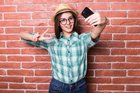 Beautiful young dark-haired girl in casual clothes and hat, eyeglasses, posing, smiling, making selfie, standing against brick wallの写真素材