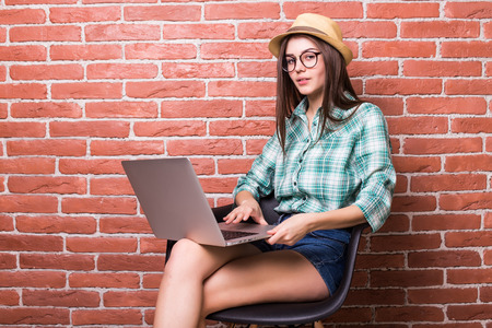 Beautiful young woman on chair in casual wear is using a laptop and smiling of tired work,  hands up, sitting against red brick wallの写真素材