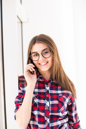 Smiling girl in casual clothes standing against window with her mirror and using mobile phone.の写真素材
