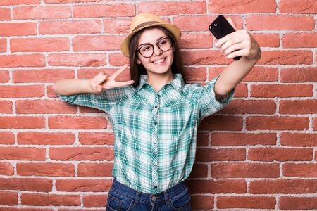 Beautiful young dark-haired girl in casual clothes and hat, eyeglasses, posing, smiling, making selfie, standing against brick wallの写真素材