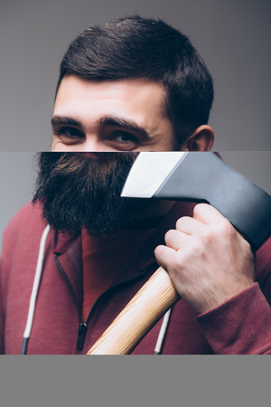 Shaving with axe. Portrait of Confident young bearded man carrying a big axe on shoulder and looking at camera while standing against grey backgroundの写真素材