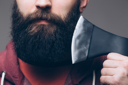 Close up of beard Confident young bearded man carrying a big axe on shoulder and looking at camera while standing against grey backgroundの写真素材