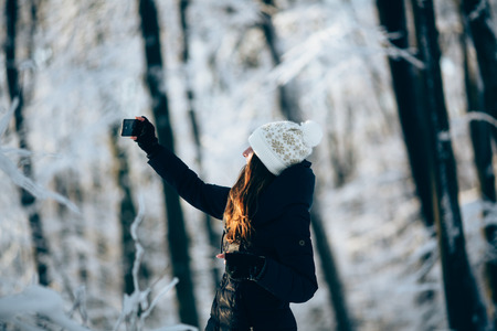 Girl Wearing Warm Winter Clothes And Hat  outdors in the forest taking photo with phone (selfie)の写真素材