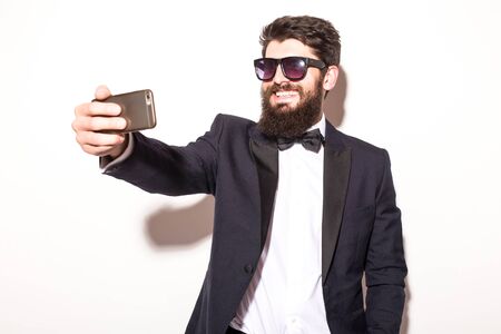 handsome young hipster man in suit and sunglasses making selfie from hand and smiling while standing against grey backgroundの写真素材