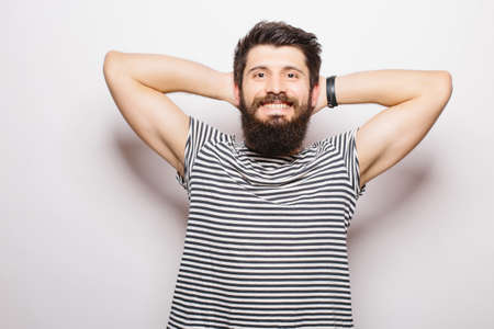 Handsome young man with beard holding hands behind head and smiling while standing against white backgroundの写真素材