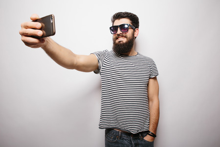 Smiling happy hipster man in sun glasses with beard standing on white isolated background and taking selfie with mobile phone.の写真素材