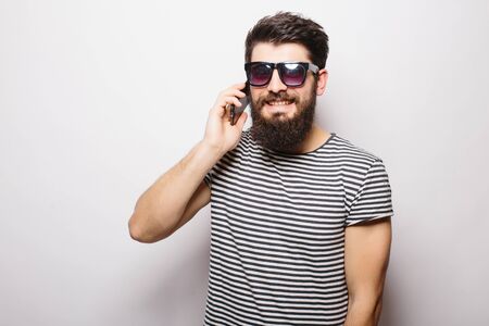 Handsome happy man with beard in sunglasses talking on the phone against white backgroundの写真素材