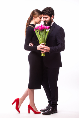Portrait of young family couple in love with bouquet of lila tulips posing dressed in classic clothes on white backround. Man with beard in Suit, Woman in black Dress. The concept of Spring holidayの写真素材