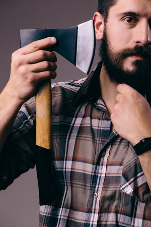 Shaving with axe. Confident young bearded man carrying a big axe on shoulder and looking at camera while standing against dark backgroundの写真素材