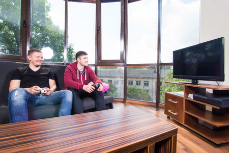 Two young happy men playing video games while sitting on sofaの写真素材