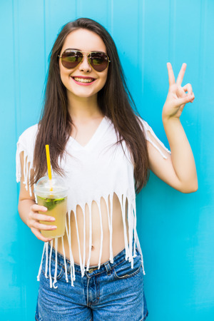 Fashion pretty woman with lemonade or fresh  in sunglasses and hat over blue colorful backgroundの写真素材