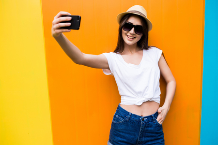 Fashion happy woman makes self portrait on smartphone  over city color wall backgroundの写真素材