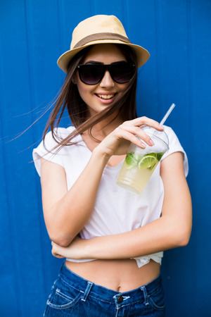 Fashion pretty woman with lemonade or fresh  in sunglasses and hat over blue colorful backgroundの写真素材
