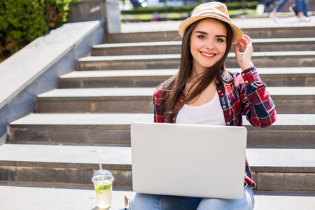 Portrait of a happy young woman sitting on the city stairs and using laptop computer outdoors look at cameraの写真素材