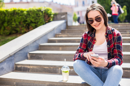 Happy young woman with a fresh cup sitting on the stairs and using her smartphone for communication.の写真素材