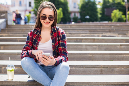 Happy young woman with a fresh cup sitting on the stairs and using her smartphone for communication.の写真素材