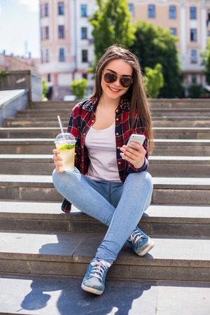 Happy young woman with a fresh cup sitting on the stairs and using her smartphone for communication.の写真素材