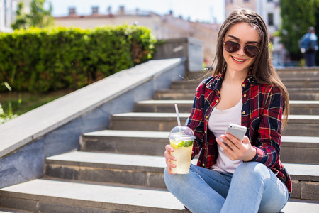 young woman with a limonade cup sitting on the stairs and text on her smartphoneの写真素材