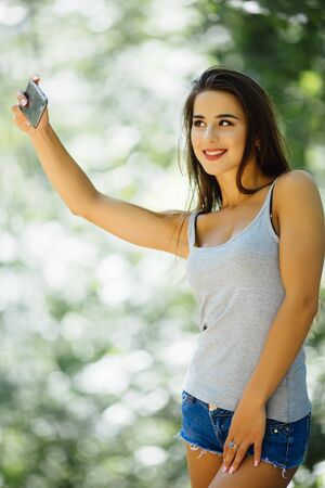 Portrait of a beautiful young woman selfie in the park with a smartphoneの写真素材