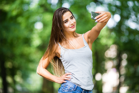 Outdoor portrait of beautiful girl taking a selfie with mobile phone in city park.の写真素材
