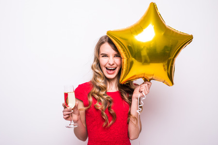 Pretty young woman in red dress with gold star shaped balloon smiling and drinking champagne over white backgroundの写真素材