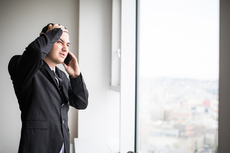 Young business man standing next to large windows of his top floor office, looking at the view of the city while take bad news from his speak on mobile phoneの写真素材