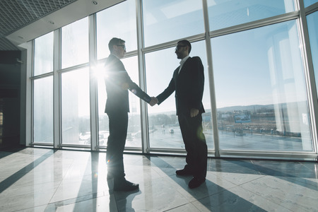 Full length side view of businessmen shaking hands in against panoramic windowsの写真素材