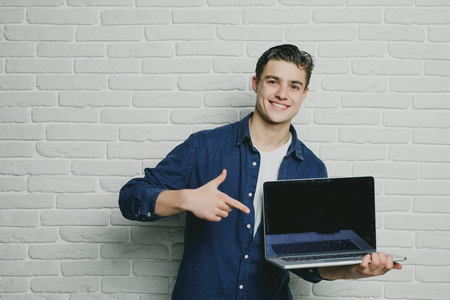 Young handsome man point the finger at a blank screen laptop, looking at the camera, happy smile, against the background of a brick wallの写真素材