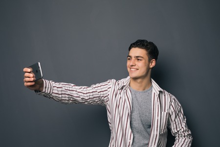 Cheerful selfie. Cheerful young man in shirt holding mobile phone and making photo of himself while standing against grey backgroundの写真素材