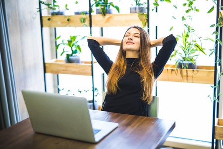 Happy young businesswoman holding hands behind head and smiling while sitting at her working placeの写真素材