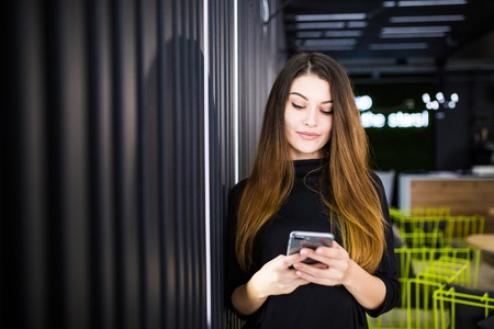 Beautiful woman chatting on mobile phone, young female in good mood reading text message on cell telephone in officeの写真素材