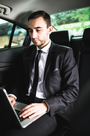 Always in touch. Handsome young businessman working on his laptop while sitting in the carの写真素材