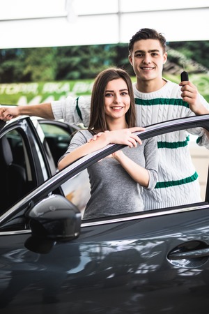 Beautiful young couple is smiling and looking at camera while leaning on their new car in a motor...