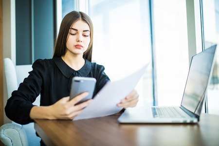 Young business woman sitting at table. Woman signs documents, hold graph and using smartphone. On table is laptop and tablet computer.の写真素材