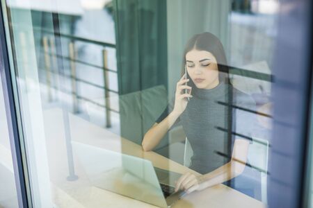Young woman talking on phone and using tablet computer through the window in officeの写真素材