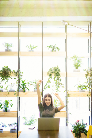 Young businesswoman sitting at an office desk in front of laptop with her hands up, looking forward to business success.の写真素材