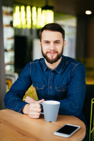 Handsome young man enjoying coffee in cafe while sitting at the table with phone laying near him in shopの写真素材