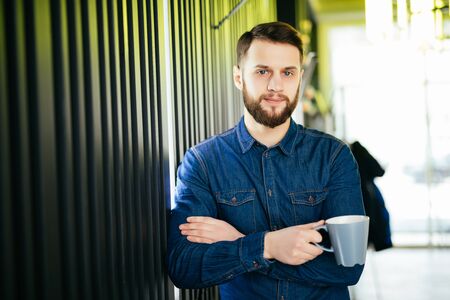Handsome businessman is holding a cup and looking away while standing in officeの写真素材
