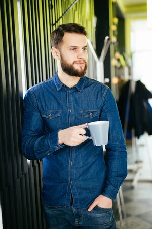 Handsome businessman is holding a cup and looking away while standing in officeの写真素材