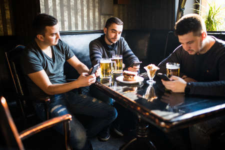 people, men, leisure, friendship and technology concept - male friends with smartphones drinking beer at bar or pubの写真素材