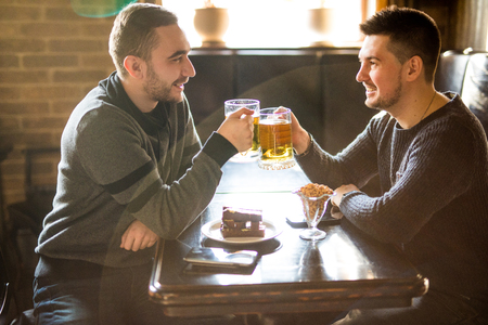 Two cheerful young men toasting with beer and smiling while sitting together in pubの写真素材