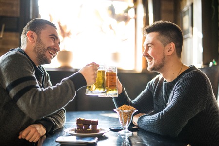 Two cheerful young men toasting with beer and smiling while sitting together in pubの写真素材