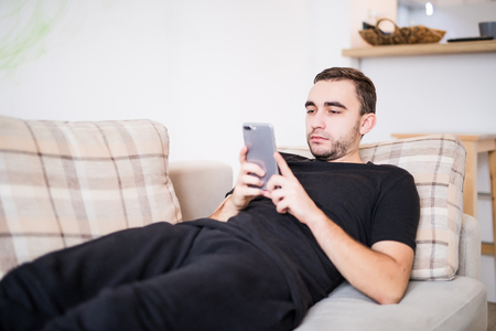 Happy to be at home. Smiling bearded man holding mobile phone and looking on it while laying on sofaの写真素材
