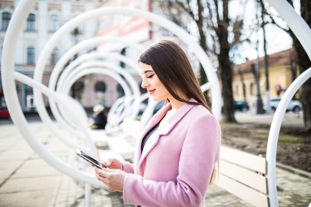 Side view of thoughtful young woman in pink jacket typing something on mobile phone in the benchの写真素材
