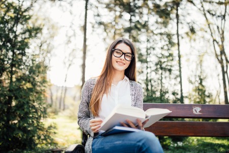 young woman sitting on bench in park, reading book, smiling and looking into the cameraの写真素材