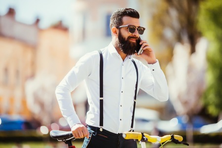 Young bearded man talking on the mobile phone and smiling while sitting near his bicycle outdoorsの写真素材